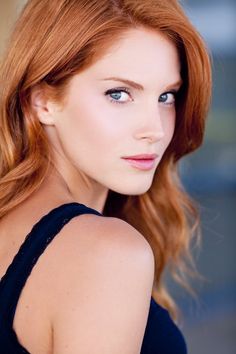 a woman with long red hair and blue eyes looks into the camera while wearing a black dress