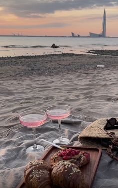 two glasses of wine are sitting on the beach with bread and croissants