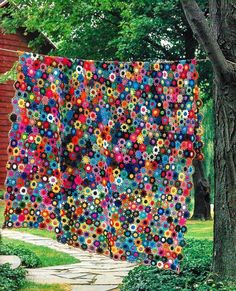 a large quilt hanging from the side of a tree in front of a house with flowers on it