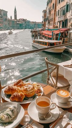 a table with coffee, croissants and bread on it next to the water