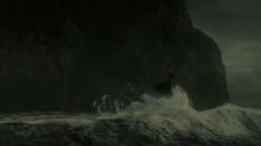 a boat in the ocean near a large rock formation with waves crashing against it and dark clouds overhead