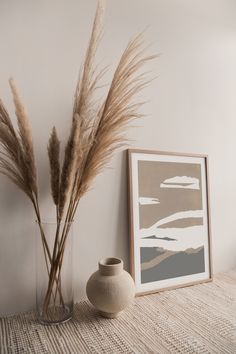 a vase with some dry grass in it next to a painting and a plant on a table