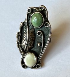 "an amazing and amazingly tall southwestern ring, artisan-made. top stone looks like a green turquoise, bottom one is a mother-of-pearl. beautiful feather and details, nice condition. marked with a stamping on the back. has a fair amount of patina but not much in the way of wear. mother-of-pearl shows some surface wear. size: 5.75 height of face: 1 and 15/16\" width of face: a bit over 13/16\" width of inner band: a bit over 1/16\"" Vintage Green Opal Ring, Vintage Green Turquoise Ring With Inlay, Vintage Green Turquoise Cabochon Ring, Handmade Southwestern Green Rings, Unique Green Inlay Rings, Bohemian Green Rings With Natural Stones, Green Bohemian Jewelry With Inlay, Bohemian Green Jewelry With Inlay, Bohemian Green Inlay Jewelry