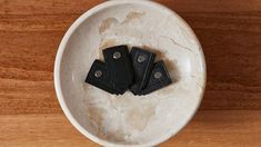 three pieces of black leather sitting in a white bowl on a wooden surface with metal rivets