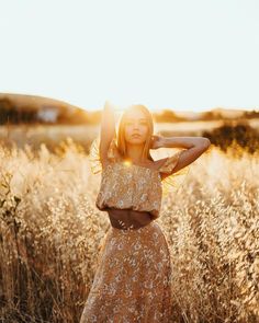 a woman standing in tall grass with her arms behind her head, wearing a crop top and skirt