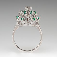 This domed cluster cocktail ring features seven (7) round brilliant cut diamonds that are prong set. The diamonds are accented with twelve (12) round mixed cut emeralds that are also prong set. The ring measures 15.2mm at the top, rises 11.7mm above the finger, tapering to 1.7mm wide and 1.3mm thick at the base of the shank. It is currently a size 6.75. White Gold Emerald Ring With Brilliant Cut Cluster, White Gold Cluster Emerald Ring With Brilliant Cut, White Gold Emerald Cluster Ring, Emerald Cluster Diamond Ring Fine Jewelry, Emerald Cluster Ring In White Gold, Emerald Cluster Ring With Brilliant Cut Diamonds, Emerald Cluster Ring With Brilliant Cut, Formal Cluster Brilliant Cut Emerald Ring, Formal Cluster Emerald Ring