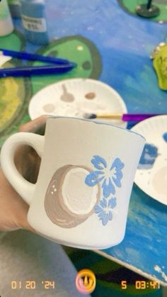 a person holding a coffee cup in front of paintbrushes and paper plates on a table