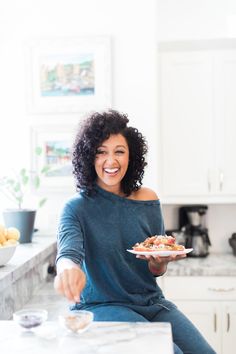 a woman sitting at a kitchen counter with a plate of food in her hand and smiling