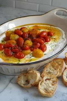 a white dish filled with bread and tomatoes on top of a marble counter next to garlic bread