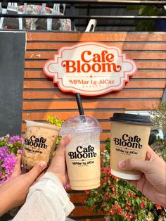 two people holding up drinks in front of a cafe bloom sign and flower potted plant