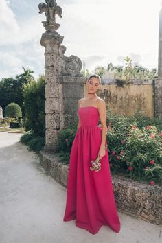 a woman in a long pink dress standing next to a stone wall and flower garden