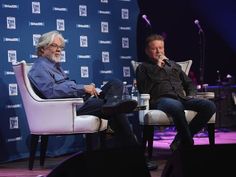 two men sitting in chairs talking on stage with microphones to the side and blue backdrop behind them