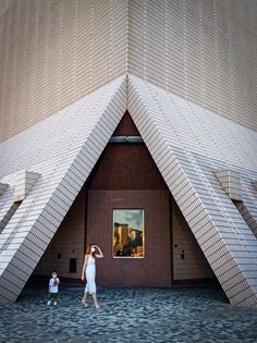 two people walking in front of a building with large triangular shaped windows and brick walls