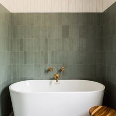 a white bath tub sitting in a bathroom next to a wooden stool and tiled walls