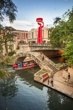 a red sculpture on the side of a river next to a bridge and people walking