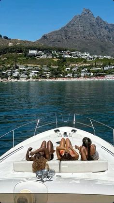 two women lounging on the back of a boat in the water with mountains in the background