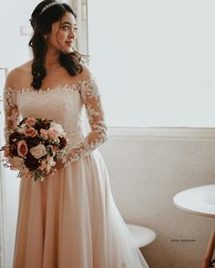 a woman in a wedding dress is standing near a window and holding a flower bouquet