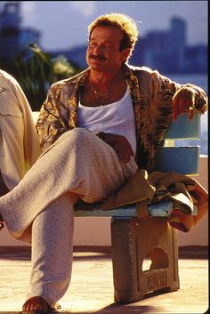 a man sitting on top of a wooden bench next to another man wearing white pants