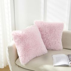 two pink pillows sitting on top of a white couch next to a book and window