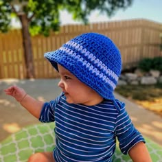 a baby wearing a blue crocheted hat sitting on a blanket in the yard