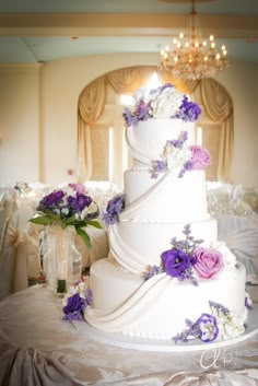 a three tiered wedding cake with purple flowers on the top and bottom, sitting on a table