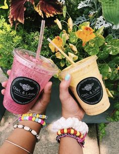 two people holding up drinks in front of some plants and flowers with their hands together