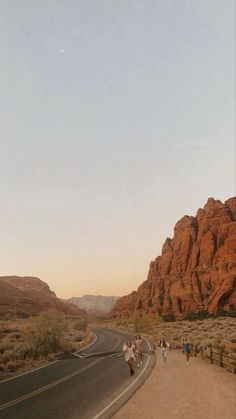 people are walking down the road in front of some red rock formations and desert like terrain