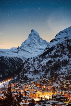 a snowy mountain town at night with the lights on and snow covered mountains in the background