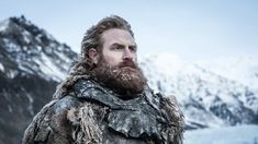 a bearded man with long hair and beard standing in front of snow covered mountains, looking off into the distance