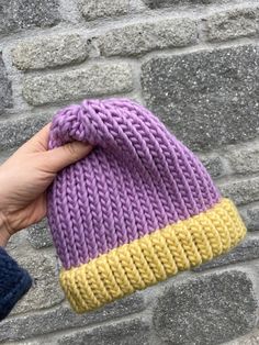 a hand holding a purple and yellow knitted hat on top of a brick wall