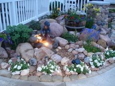 a fire pit surrounded by rocks and flowers in front of a white picket fenced porch