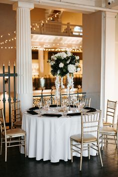 the table is set with white flowers and black plates on it, along with silver chairs