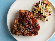 a white plate topped with meat and coleslaw next to a salad on a blue table