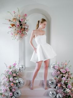 a woman in a short white dress standing next to flowers