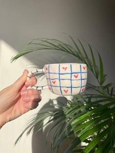 a hand holding a coffee cup with hearts painted on the outside and inside, in front of a potted plant