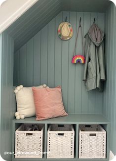 a bench with baskets and pillows on it in a room that has blue walls, green paneling, and two coats hanging from the wall