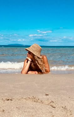 a woman laying on top of a sandy beach next to the ocean wearing a hat