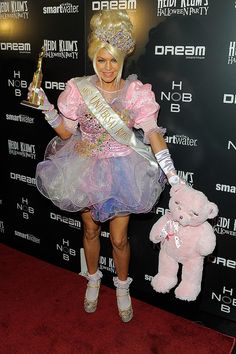 a woman in a pink dress holding a teddy bear and wearing a tiara while standing on a red carpet