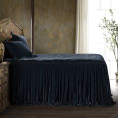 a bed with black sheets and pillows in a room next to a dresser, potted plant and window