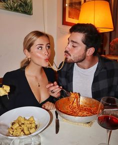 a man and woman sitting at a table with spaghetti in front of their mouths as they eat pasta