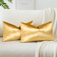 two gold pillows sitting on top of a white couch next to a green potted plant