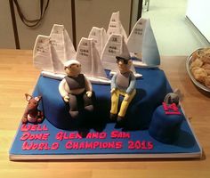 a cake with two men sitting on top of it and some cookies in the background