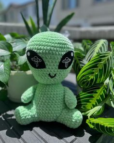 a crocheted alien sitting next to some plants