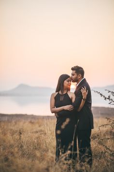 a man and woman standing in tall grass with the sun setting on the water behind them