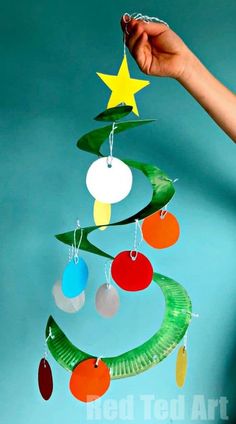 a hand holding a paper plate christmas tree mobile with ornaments hanging from it's sides