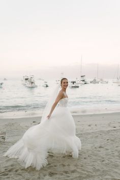 a woman in a wedding dress is walking on the beach with boats in the background