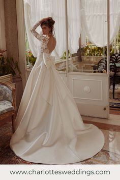 a woman in a wedding dress standing by a window with her hands on her head