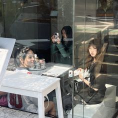 two women sitting at a table in front of a glass window, taking pictures with their cell phones