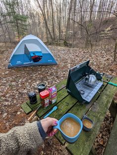 someone is camping in the woods with their laptop and cup of coffee on a picnic table