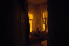 a person sitting on a couch in a dark room with curtains and sunlight coming through the window